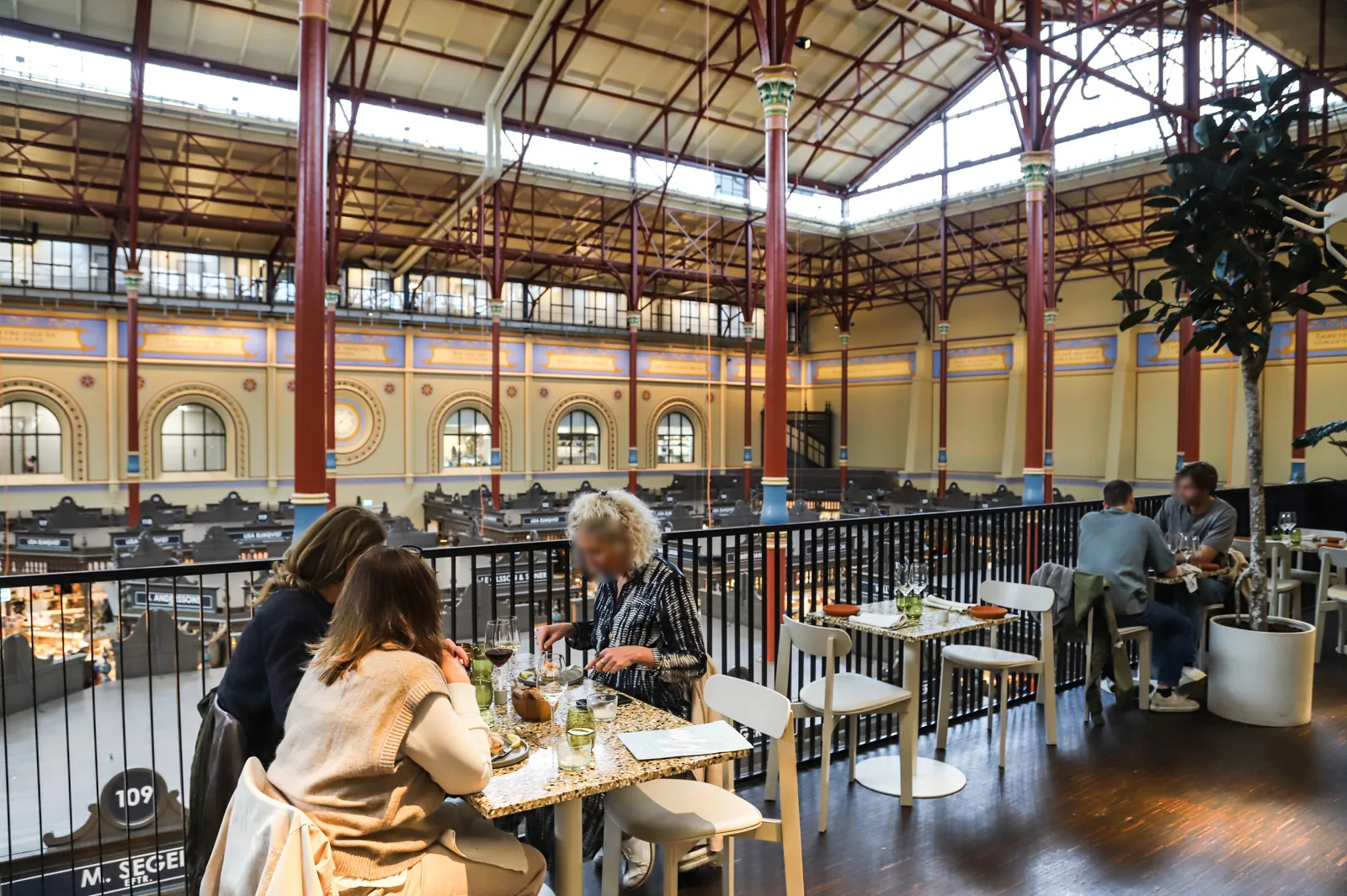 diners enjoy meals at tables in a historic industrial market hall with arched windows., östermalms food hall, stockholm