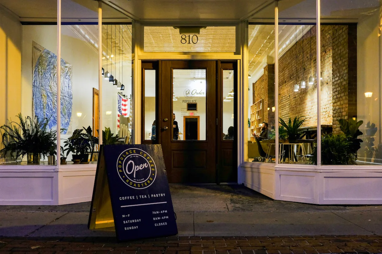 modern coffee shop storefront at night with illuminated yellow brick facade and plants., populace coffee, bay city