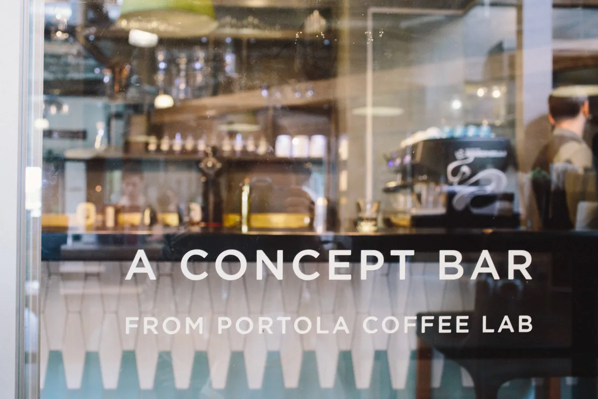glass storefront displaying "a concept bar from portola coffee lab" signage with interior café setup visible., portola coffee