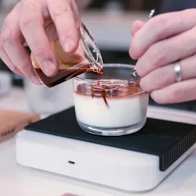 person pouring rose liquid into layered dessert in glass cup., ratio coffee (no food), crows nest