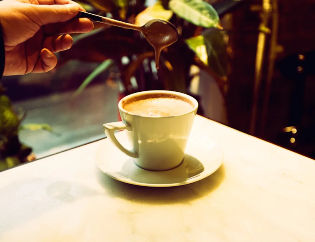 hand stirring coffee cup with saucer on white table, green plants visible background., restaurant gran delicato, helsinki