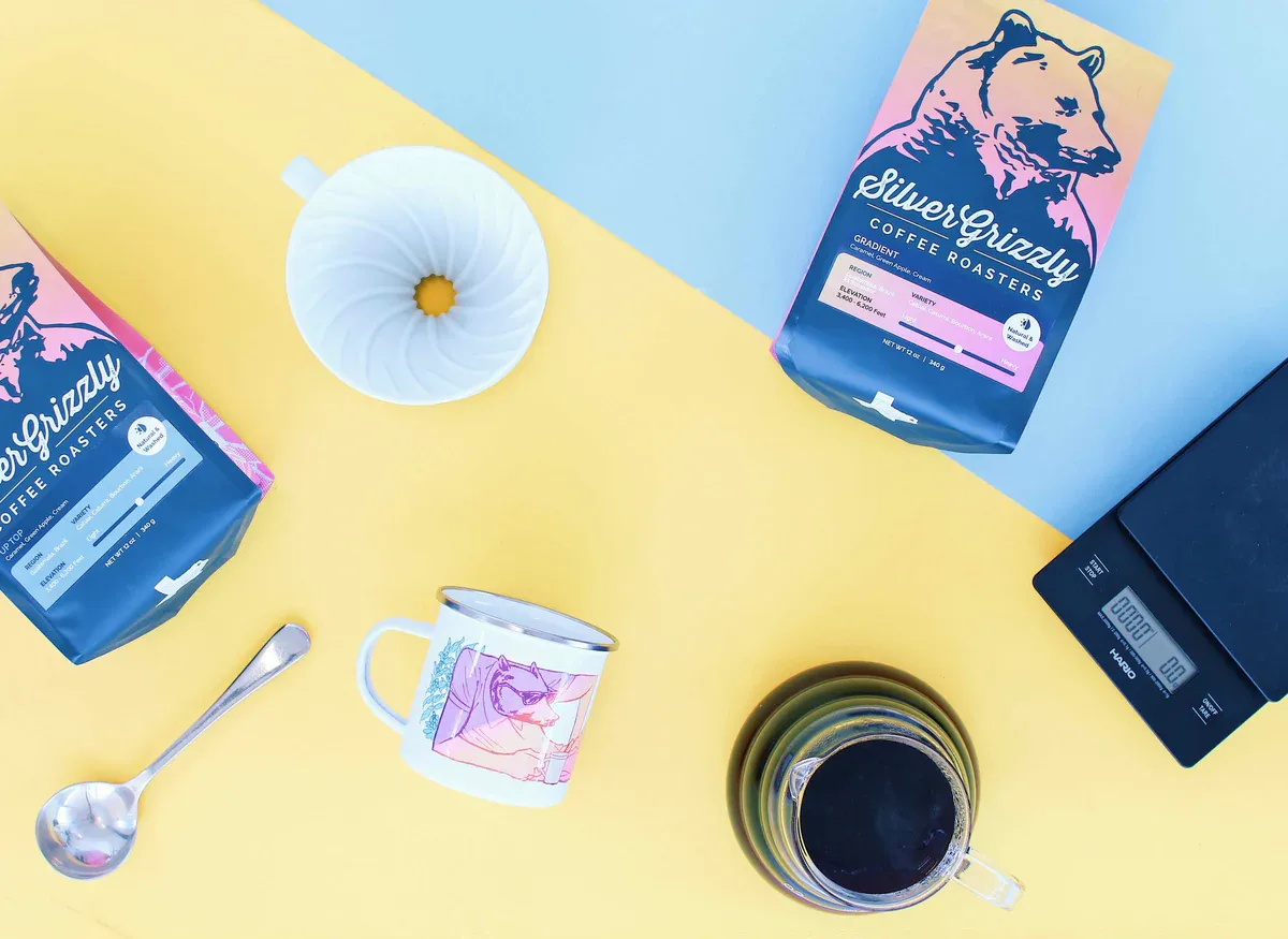 silvergrizzly coffee bags, scale, mug, spoon, flower, and brewed coffee arranged on yellow surface., silver grizzly coffee ro