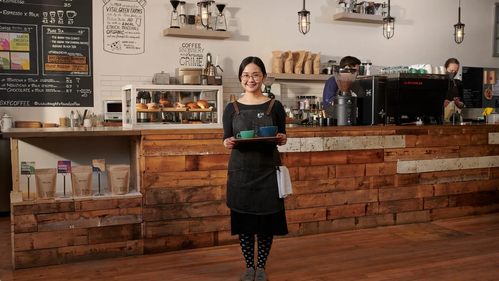 barista in black apron holding coffee cups at wooden counter cafe., sought and found coffee roasters, calgary