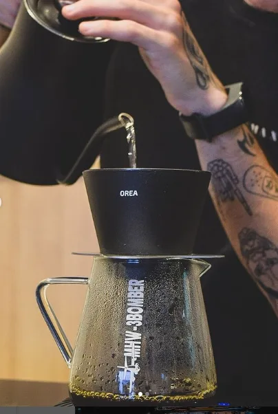 person pouring hot water into black orea dripper over glass carafe with filter., studio grão coffee roasters, brasília