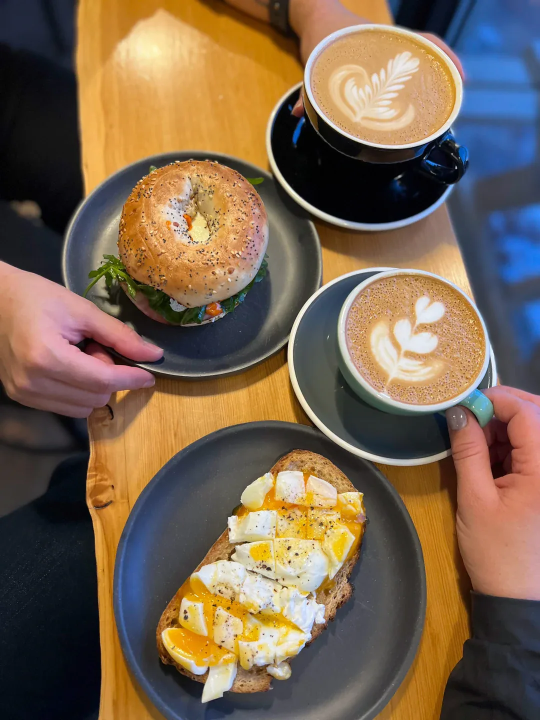 two lattes with latte art, seeded bagel, and poached egg toast on wooden table., the colombian coffee bar & roastery- glenora