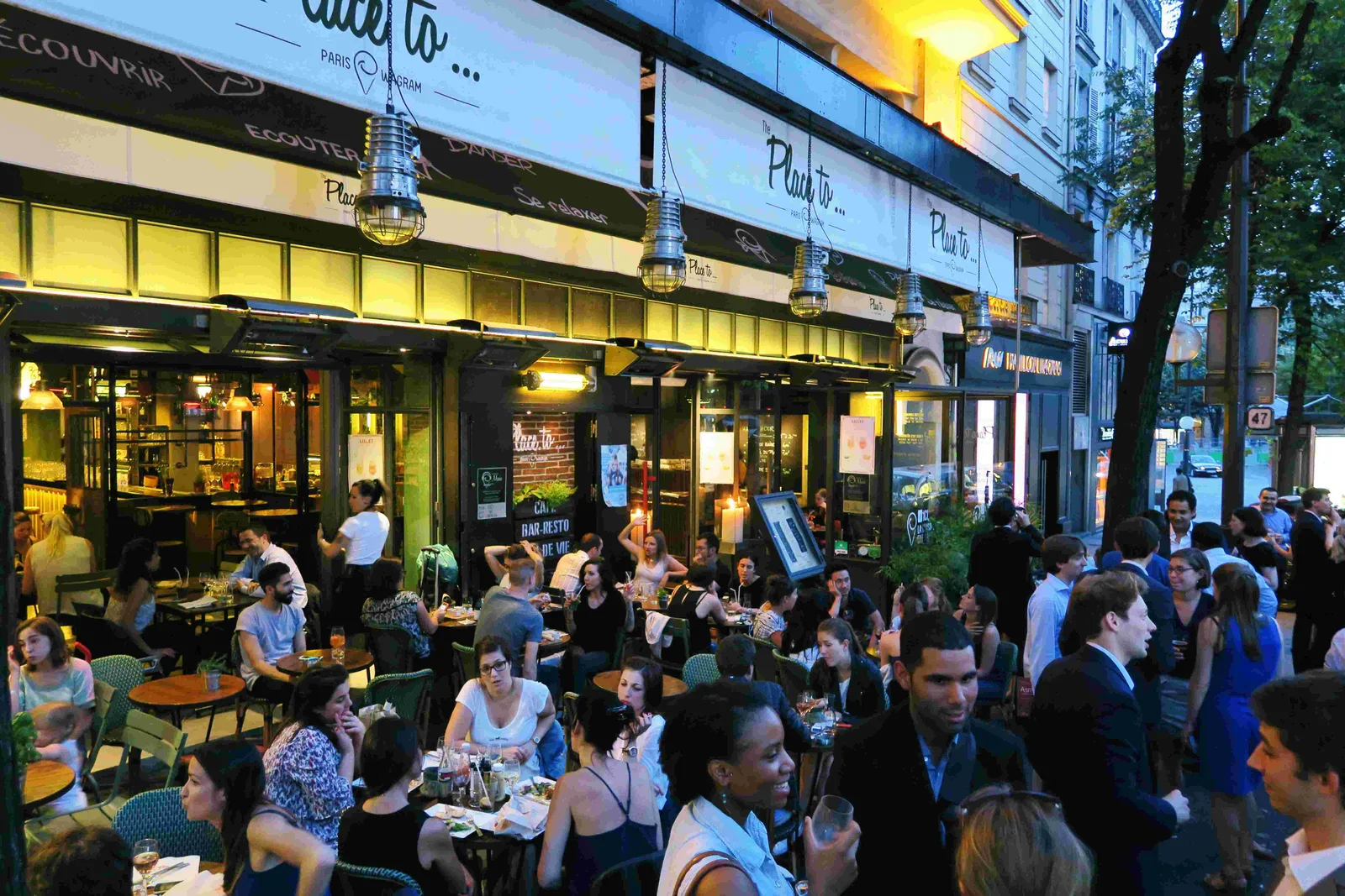 crowded outdoor parisian café with diners, illuminated storefront, evening atmosphere., the place to...wagram - restaurant pu