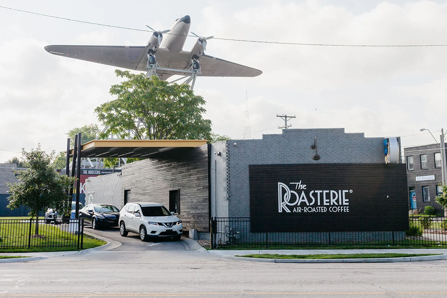 vintage airplane mounted above coffee roastery building with parked cars below., the roasterie, kansas