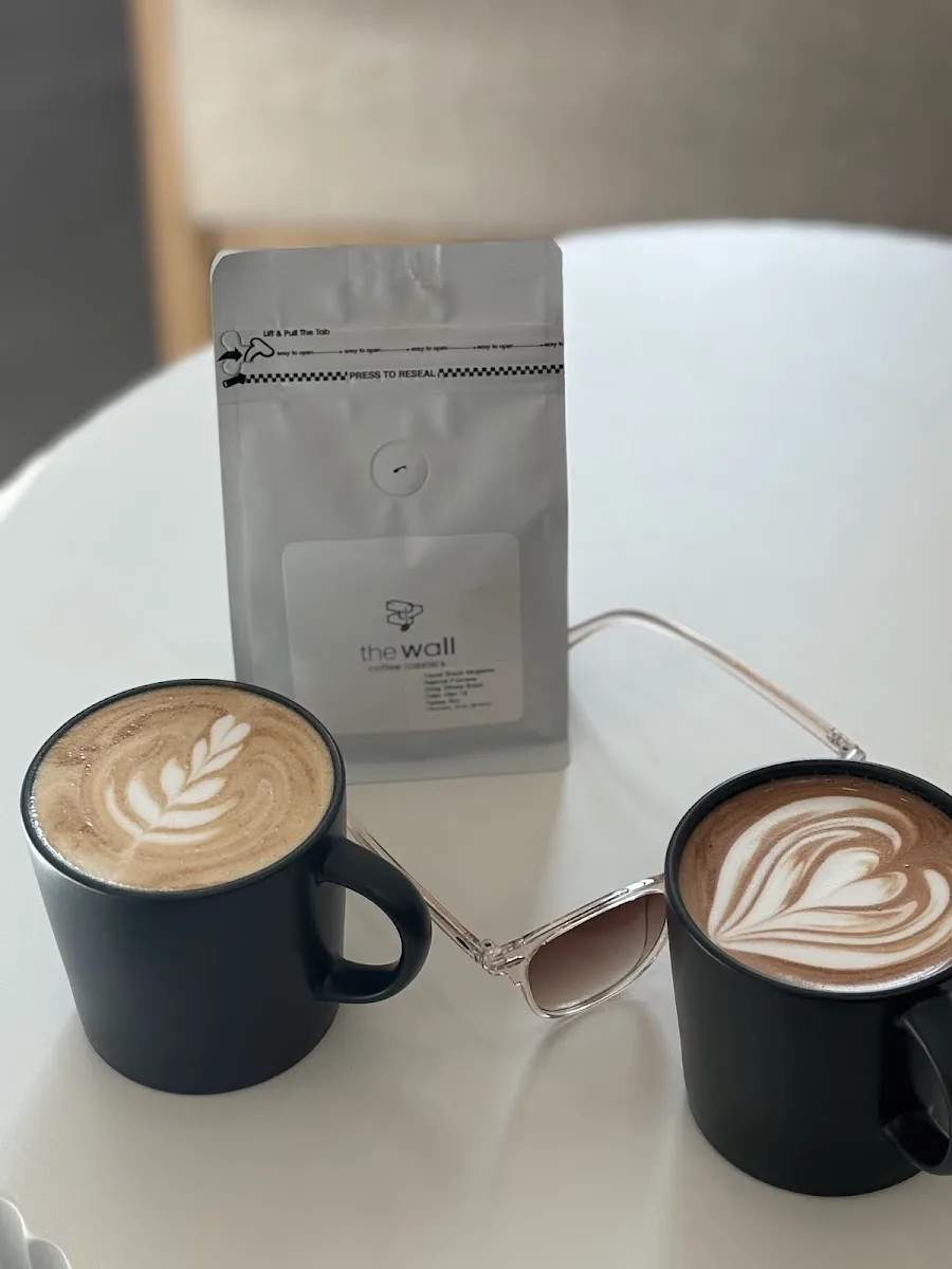 two black mugs with latte art, coffee bag labeled "the wall," and sunglasses on white table., the wall coffee roasters, aches