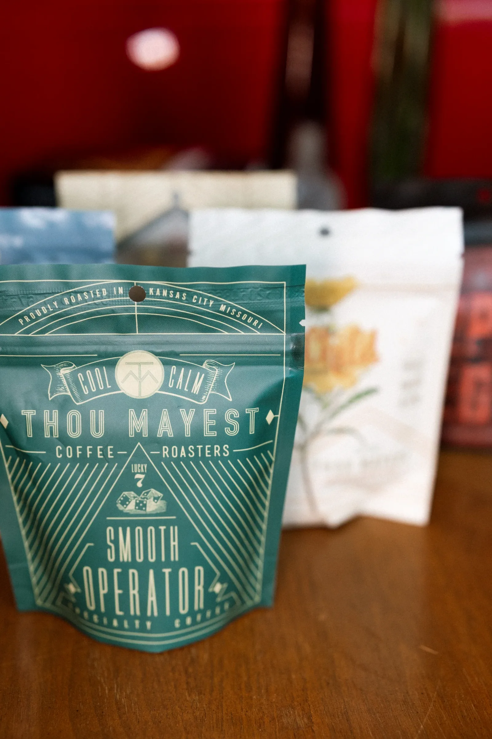 teal coffee bag labeled "thou mayest" from kansas city with blurred packages behind., thou mayest coffee roasters, kansas cit
