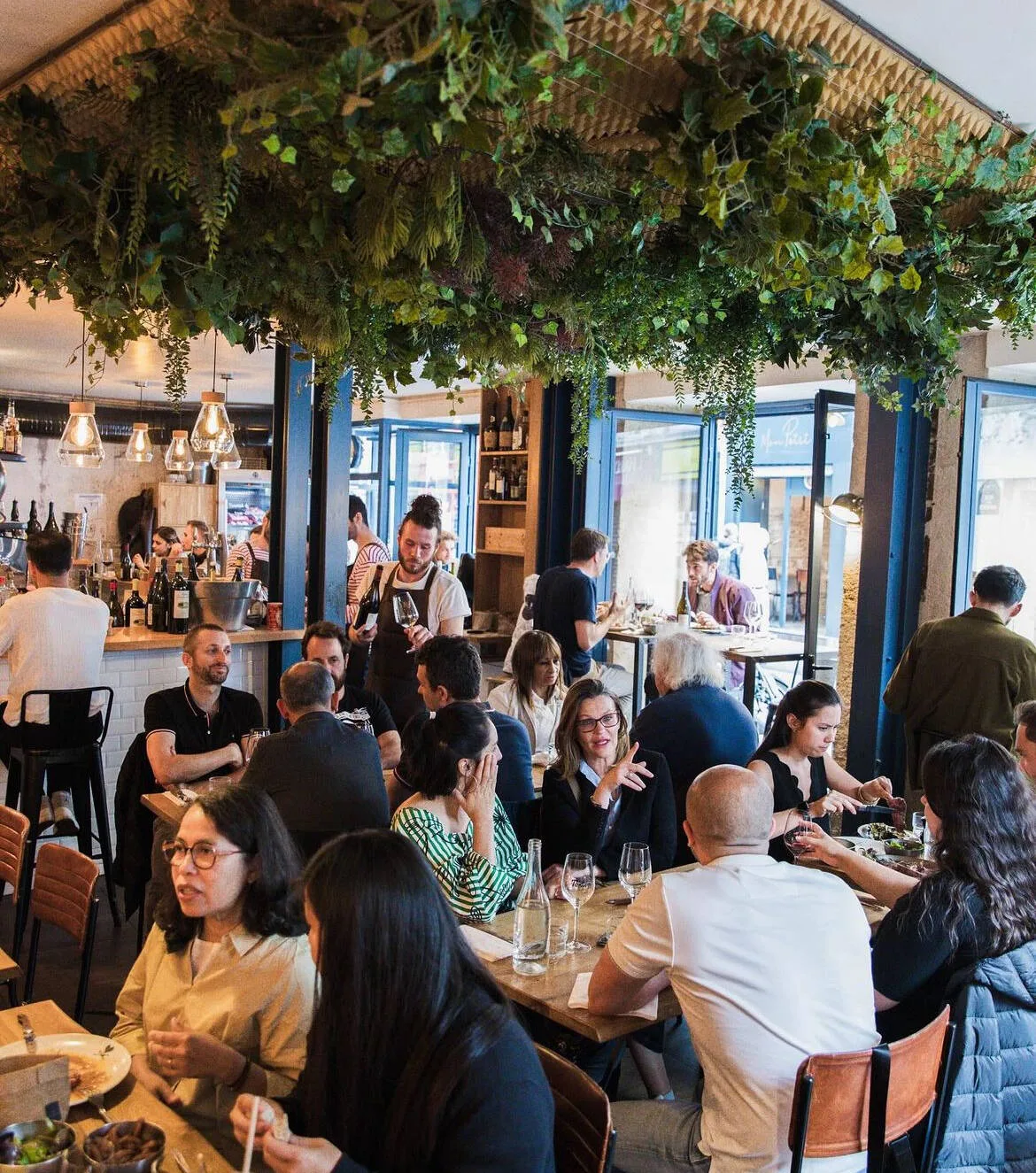 bustling restaurant interior with diners, bar counter, and hanging plants overhead., tonton des dames, paris