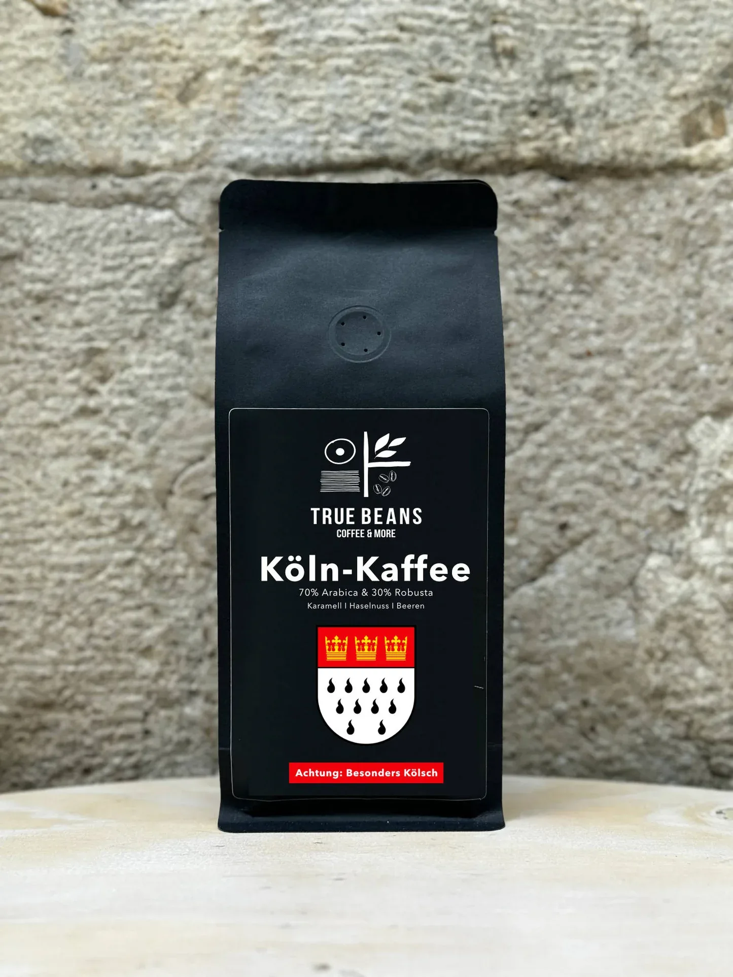 black coffee bag featuring cologne coat of arms against textured brick wall., truebeans specialty coffee - werksverkauf & sem