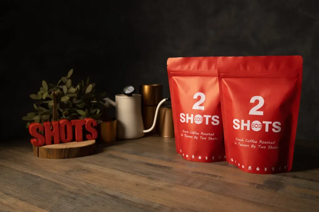 two red coffee pouches beside brass kettle and potted plant on wooden surface., two shots 圖夏次咖啡 頂溪店, taipei