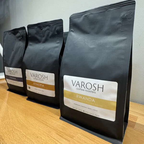 three black varosh coffee bags labeled ethiopia, rwanda, and partially visible third variety on wooden surface., varosh coffe