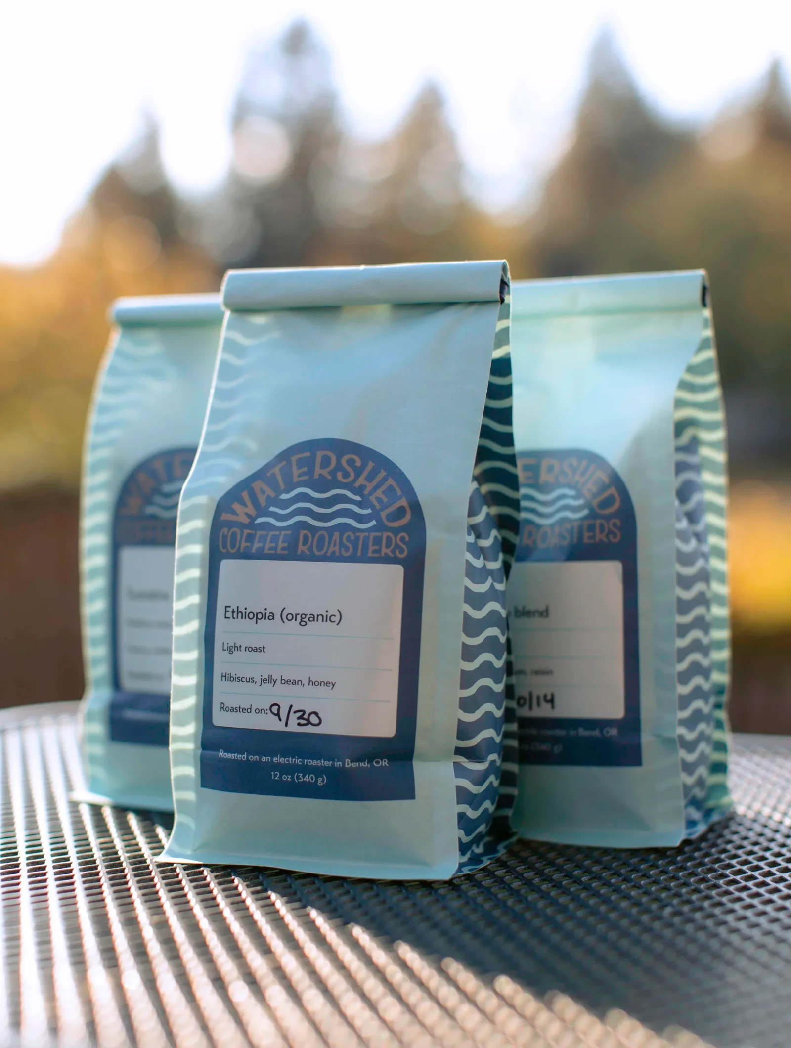 watershed coffee roasters bags displayed outdoors with evergreen trees blurred in background., watershed coffee roasters, ben