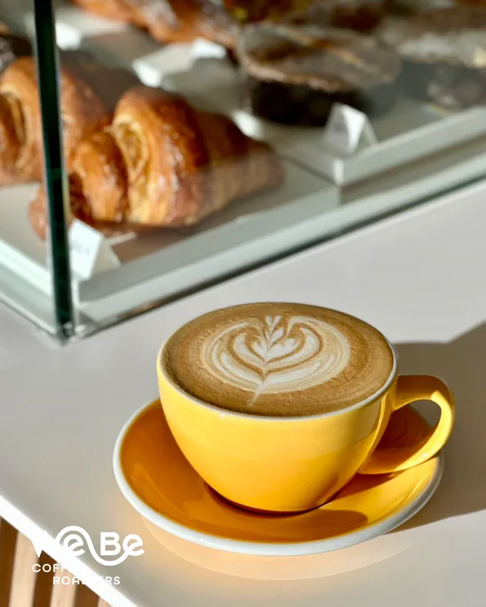 yellow cup with latte art on saucer, pastries visible in background display case., webe coffee roasters, phoenix