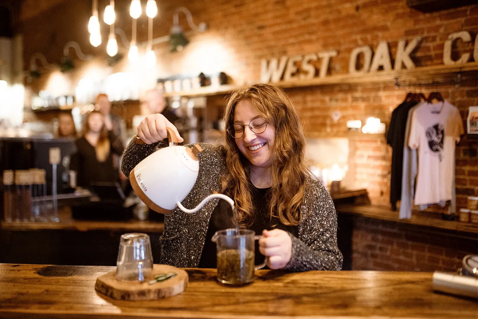 woman pouring hot water into glass at wooden bar counter in brick venue., west oak coffee bar, texas