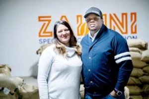 two people posing together in front of an orange "zabbini" branded sign., zabuni coffee, nebraska